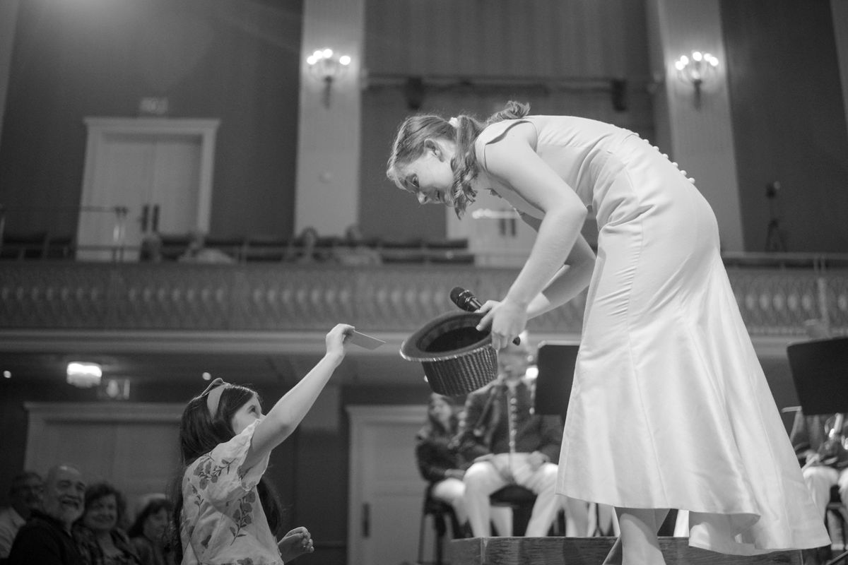 A child draws a note from a hat held out by Alma Deutscher on a concert stage