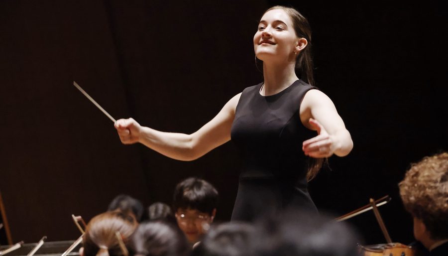 Alma Deutscher conducting, smiling, with musicians of the orchestra in the foreground